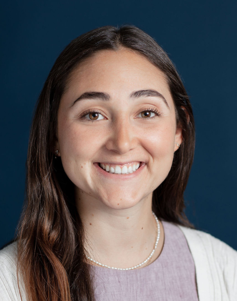 A woman with long brown hair, wearing a light purple top and cream cardigan, smiles in front of a solid dark blue background.